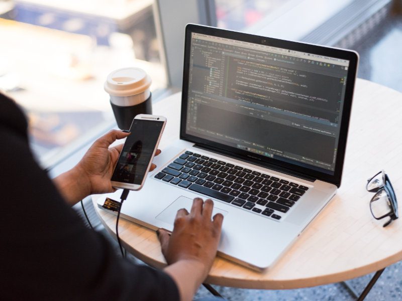 A person holds a phone in one hand and has their other hand on a MacBook Pro, which displays a code editor