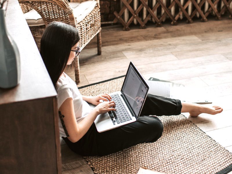 A woman sits on a mat on the floor and types on a MacBook Pro